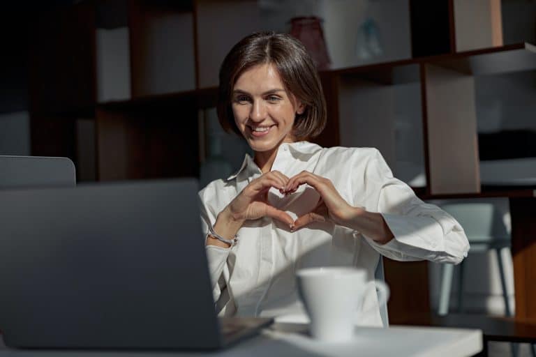 Businesswoman making video call to friends and showing sign love while working in modern office